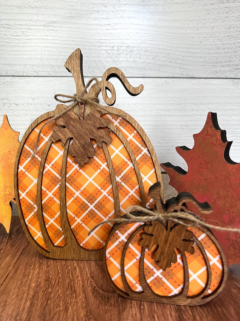 May include: Two wooden pumpkin-shaped decorations featuring a cutout frame design with an orange and white plaid pattern background. Each pumpkin is accented with a dark brown wooden leaf cutout and a tied piece of thin brown twine.