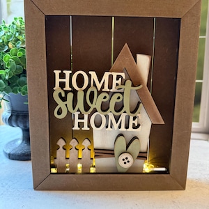 May include: A wooden framed decorative piece with the words "HOME Sweet HOME" in layered lettering. The design includes a house, fence, and heart, with small lights illuminating the interior. The frame is brown.