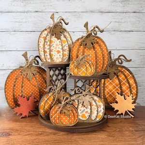 May include: A tiered wooden display stand holding multiple flat pumpkin-shaped decorations. The pumpkins feature various orange and white patterns including polka dots, plaid, and autumn leaves. Each pumpkin is topped with a wooden leaf cutout.