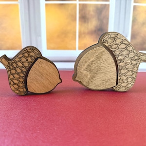 May include: Two wooden acorn-shaped ornaments of different sizes. Each piece features a smooth, light-colored wood center and a darker, laser-engraved cap with a detailed cross-hatched scale pattern. The edges are dark, highlighting the shapes.