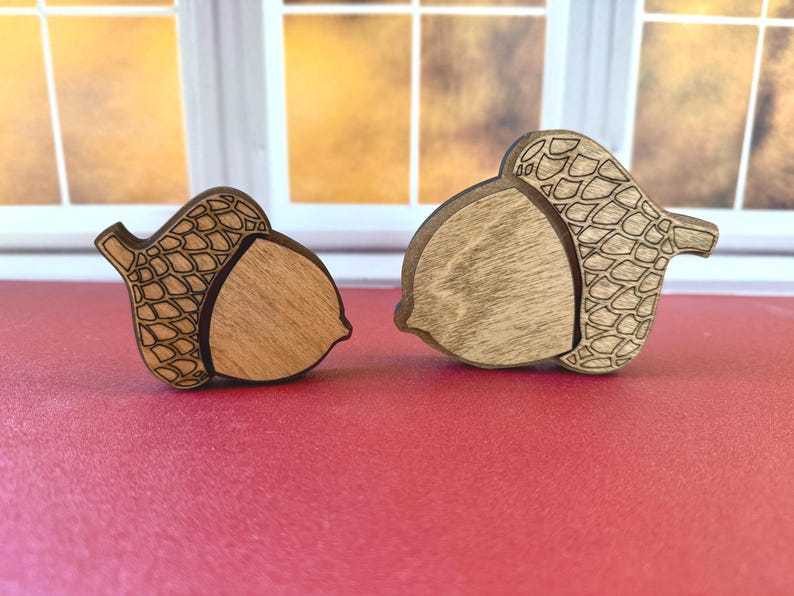 May include: Two wooden acorn-shaped ornaments of different sizes. Each piece features a smooth, light-colored wood center and a darker, laser-engraved cap with a detailed cross-hatched scale pattern. The edges are dark, highlighting the shapes.