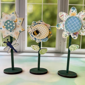 Puede incluir: Tres figuras decorativas de flores de madera con pétalos estampados y tallos verdes. Una tiene una libélula, otra una abeja y la tercera un patrón de cuadrícula. Cada flor está sobre una base circular verde.