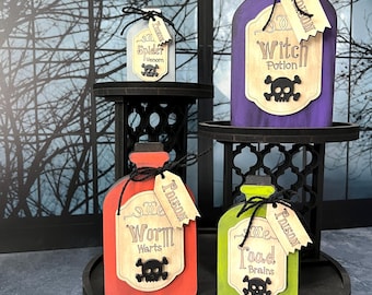Puede incluir: Cuatro botellas de poción de madera en gris, morado, naranja y verde, cada una con una etiqueta beige con texto y un icono de calavera negra. Las etiquetas dicen Spider Venom, Witch Potion, Worm Warts y Toad Brains, con pequeñas etiquetas de Poison.