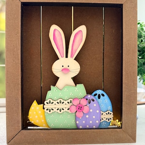 May include: Wooden Easter shadow box featuring a white bunny with pink ears emerging from a cracked green egg. Includes colorful polka-dotted eggs and a pink flower. The frame is brown.
