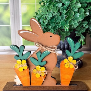May include: Wooden Easter decoration featuring a brown bunny and three orange carrots with green tops and yellow flowers. The bunny has a twine bow and is on a brown base. The background includes a green plant and a window.