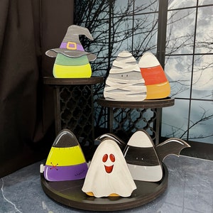 May include: A tiered wooden stand with a black finish displays a collection of Halloween-themed candy corn decorations. The decorations include a witch's hat, a mummy, a ghost, a bat, and a candy corn with a stitched-up face.