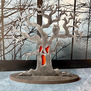 Pu&ograve; includere: Decorazione a tema Halloween a forma di albero spettrale con un gufo intagliato e un volto con interno arancione. L'albero grigio poggia su una base rotonda. Lo sfondo mostra una finestra con rami di alberi spogli.