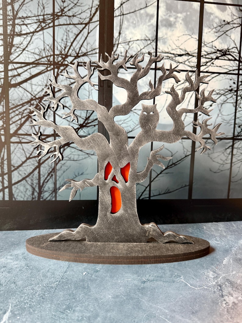 Pu&ograve; includere: Decorazione a tema Halloween a forma di albero spettrale con un gufo intagliato e un volto con interno arancione. L'albero grigio poggia su una base rotonda. Lo sfondo mostra una finestra con rami di alberi spogli.