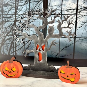Pu&ograve; includere: Decorazione di Halloween in legno con un albero spettrale, un gufo e due zucche intagliate. L'albero &egrave; grigio con un ritaglio arancione e le zucche sono arancioni con facce gialle. Lo sfondo mostra una luna e rami di alberi spogli.