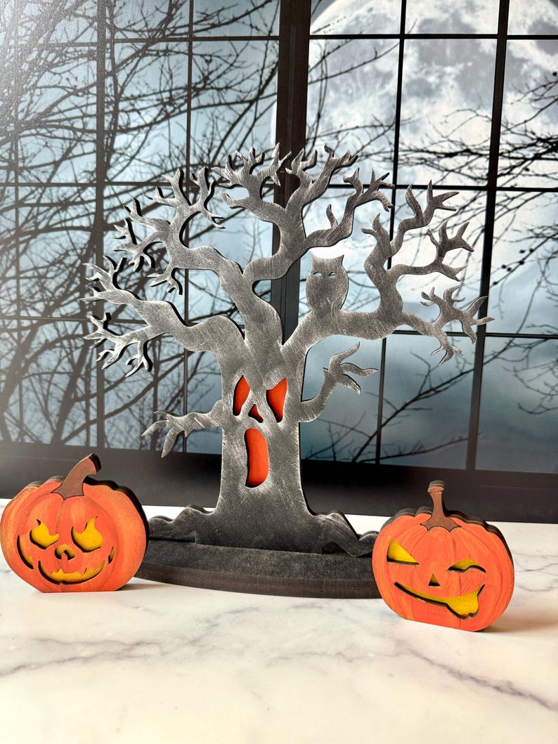 Pu&ograve; includere: Decorazione di Halloween in legno con un albero spettrale, un gufo e due zucche intagliate. L'albero &egrave; grigio con un ritaglio arancione e le zucche sono arancioni con facce gialle. Lo sfondo mostra una luna e rami di alberi spogli.