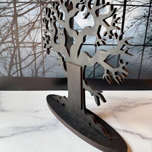 Pu&ograve; includere: Una scultura di albero in legno marrone scuro, tagliata al laser, con rami intricati e una silhouette di gufo. L'albero poggia su una base ovale. Lo sfondo presenta una finestra con rami spogli.