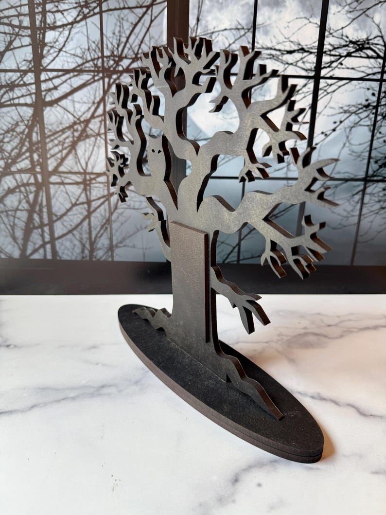 Pu&ograve; includere: Una scultura di albero in legno marrone scuro, tagliata al laser, con rami intricati e una silhouette di gufo. L'albero poggia su una base ovale. Lo sfondo presenta una finestra con rami spogli.