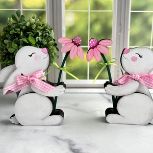 May include: Two wooden bunny figurines, each holding a pink flower with a green stem. The bunnies are white with pink cheeks, ears, and a pink and white gingham bow. The background includes a window and greenery.