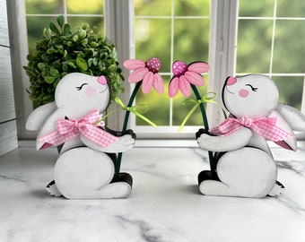 May include: Two wooden bunny figurines, each holding a pink flower with a green stem. The bunnies are white with pink cheeks, ears, and a pink and white gingham bow. The background includes a window and greenery.