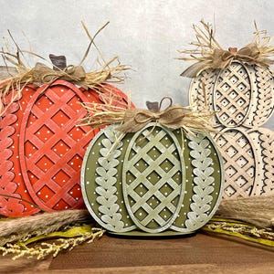 Puede incluir: Tres calabazas de madera con diferentes diseños y colores. Las calabazas están decoradas con arpillera y rafia. Las calabazas son naranja, verde y beige.