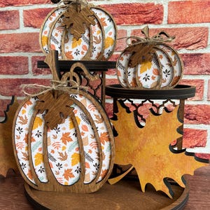 May include: A tiered wooden display stand featuring three pumpkin-shaped cutouts and one large maple leaf cutout. The pumpkins have dark brown wooden frames with white inserts printed with orange, yellow, and brown autumn leaf patterns.