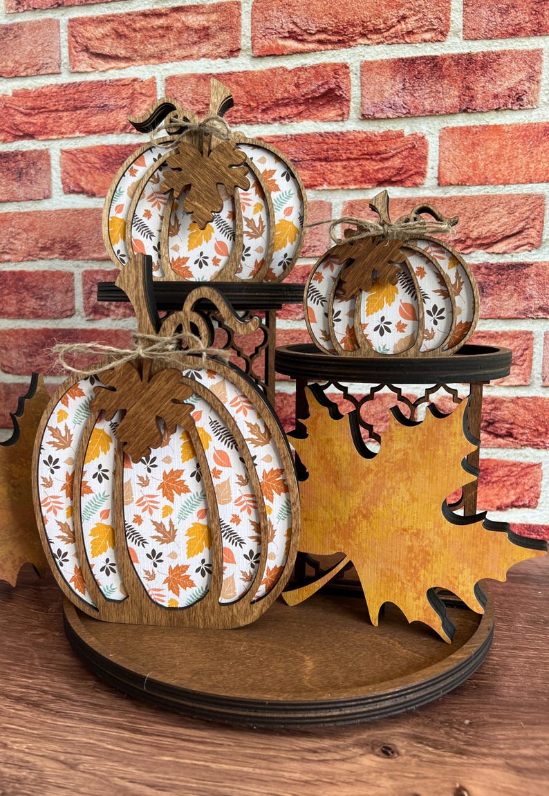 May include: A tiered wooden display stand featuring three pumpkin-shaped cutouts and one large maple leaf cutout. The pumpkins have dark brown wooden frames with white inserts printed with orange, yellow, and brown autumn leaf patterns.