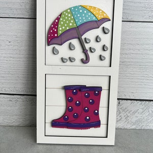 K&ouml;nnte beinhalten: Ein wei&szlig;es Holzschild mit einem bunten, gepunkteten Regenschirm und Regentropfen im oberen Teil und einem Paar rosa Gummistiefeln mit violetten Punkten im unteren Teil.