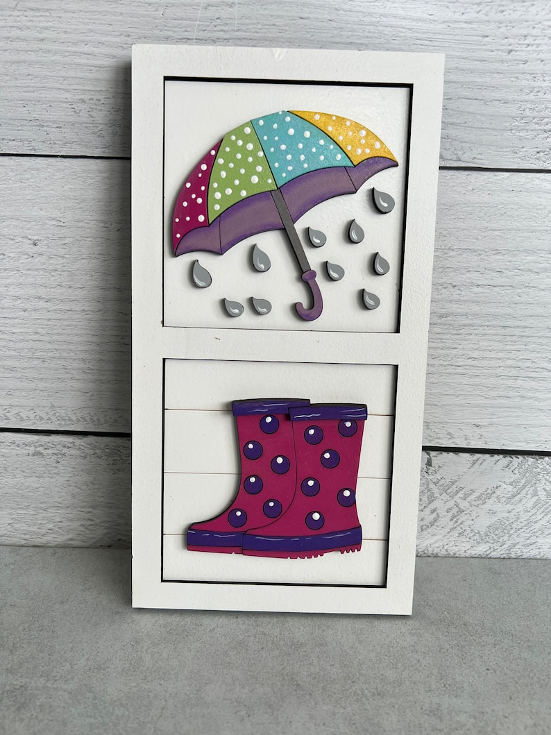 K&ouml;nnte beinhalten: Ein wei&szlig;es Holzschild mit einem bunten, gepunkteten Regenschirm und Regentropfen im oberen Teil und einem Paar rosa Gummistiefeln mit violetten Punkten im unteren Teil.