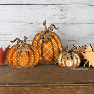 May include: A set of three wooden pumpkin decorations with brown frames and patterned inserts, including polka dot, plaid, and leaf prints. Each pumpkin features a twine bow and a wooden leaf accent. Two wooden maple leaves are placed on the sides.