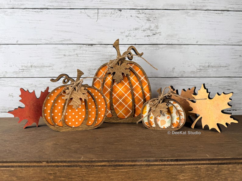 May include: A set of three wooden pumpkin decorations with brown frames and patterned inserts, including polka dot, plaid, and leaf prints. Each pumpkin features a twine bow and a wooden leaf accent. Two wooden maple leaves are placed on the sides.