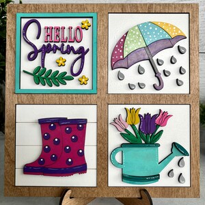K&ouml;nnte beinhalten: Ein Holzschild mit vier Quadraten, jedes mit einem anderen Fr&uuml;hlingsmotiv. Das obere linke Quadrat zeigt den Text "Hello Spring" in lila und rosa Schrift mit Blumen und einem gr&uuml;nen Blatt. Das obere rechte Quadrat zeigt einen lila und gr&uuml;nen Regenschirm mit wei&szlig;en Punkten und grauen Regentropfen. Das untere linke Quadrat zeigt ein Paar rosa Gummistiefel mit lila Punkten. Das untere rechte Quadrat zeigt eine t&uuml;rkisfarbene Gie&szlig;kanne mit einem Strau&szlig; rosa, lila und gelber Tulpen.