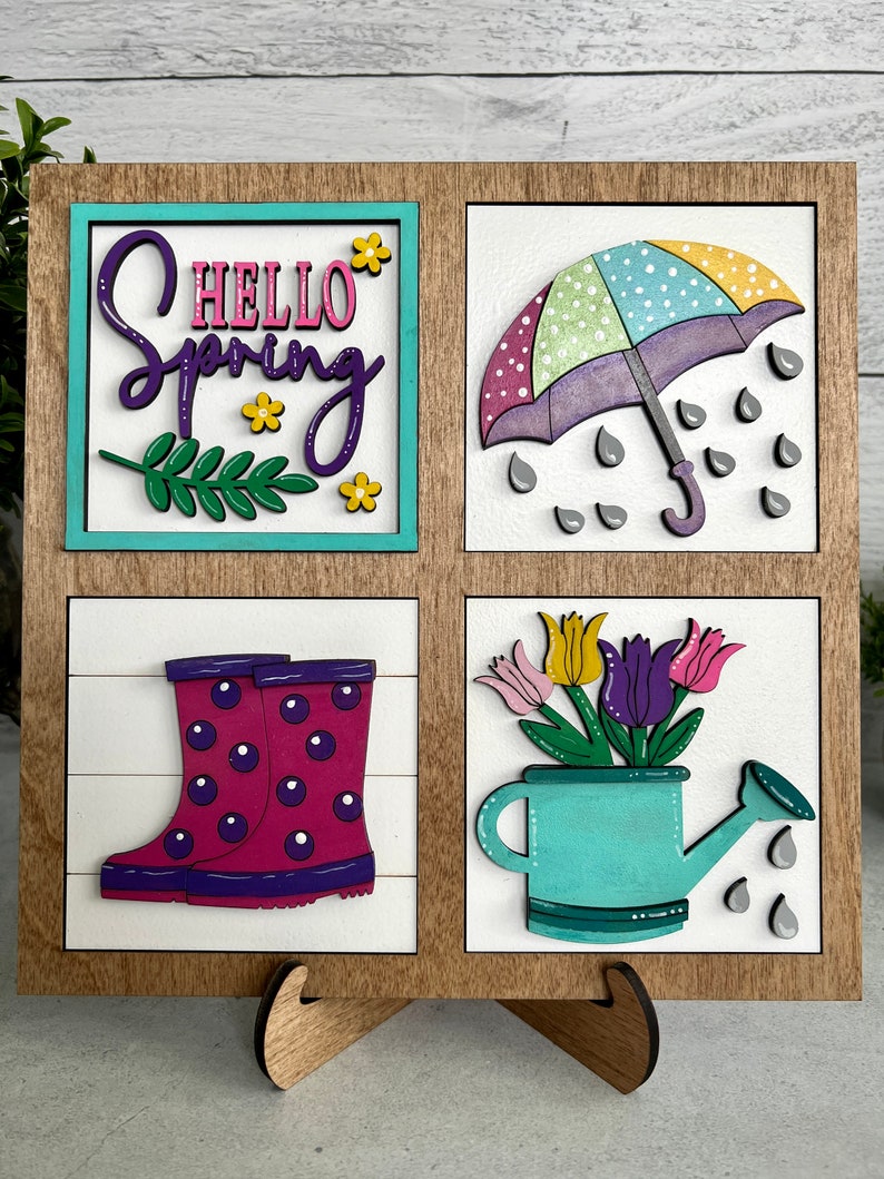 K&ouml;nnte beinhalten: Ein Holzschild mit vier Quadraten, jedes mit einem anderen Fr&uuml;hlingsmotiv. Das obere linke Quadrat zeigt den Text "Hello Spring" in lila und rosa Schrift mit Blumen und einem gr&uuml;nen Blatt. Das obere rechte Quadrat zeigt einen lila und gr&uuml;nen Regenschirm mit wei&szlig;en Punkten und grauen Regentropfen. Das untere linke Quadrat zeigt ein Paar rosa Gummistiefel mit lila Punkten. Das untere rechte Quadrat zeigt eine t&uuml;rkisfarbene Gie&szlig;kanne mit einem Strau&szlig; rosa, lila und gelber Tulpen.