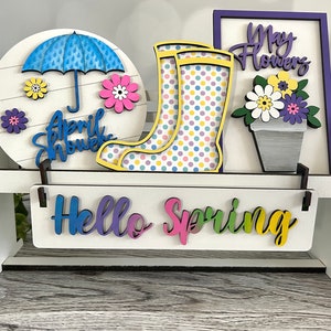 Puede incluir: Un estante de madera blanco con tres decoraciones de temática primaveral. El estante superior tiene un letrero redondo de madera con un paraguas azul, flores y el texto "April Showers". El estante del medio tiene un par de botas de lluvia amarillas con lunares de colores y un marco morado con el texto "May Flowers" y una maceta con flores. El estante inferior tiene un letrero blanco con el text "Hello Spring".