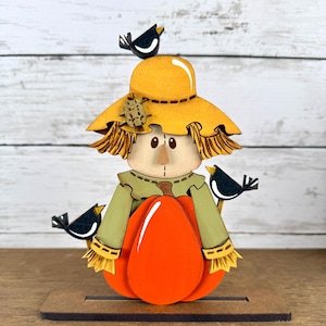 May include: A layered wooden scarecrow decoration featuring a yellow hat with a small patch, a green shirt with straw-like cuffs, and a bright orange pumpkin. Three small black birds are perched on the hat and the scarecrow's shoulders.
