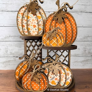 May include: A tiered wooden display stand holding five decorative pumpkins in various sizes. The pumpkins feature dark brown wooden frames with orange, white, and patterned inserts including polka dots, plaid, and autumn leaves, tied with twine.