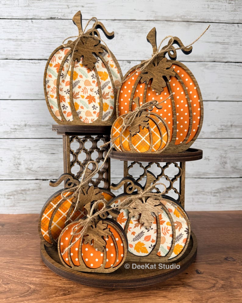 May include: A tiered wooden display stand holding five decorative pumpkins in various sizes. The pumpkins feature dark brown wooden frames with orange, white, and patterned inserts including polka dots, plaid, and autumn leaves, tied with twine.