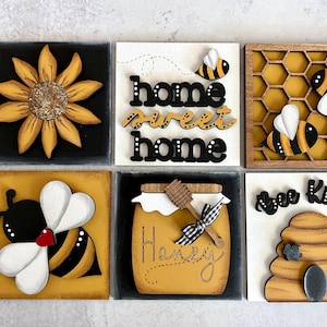May include: Six wooden signs with a bee theme. The signs feature a sunflower, a bee, a honeycomb, a jar of honey, a beehive, and the words "home sweet home" and "bee kind".