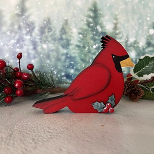 Pode incluir: Uma figura de p&aacute;ssaro cardeal vermelho com um olho e bico pretos, pousada em uma superf&iacute;cie nevada com um ramo de azevinho e bagas vermelhas.