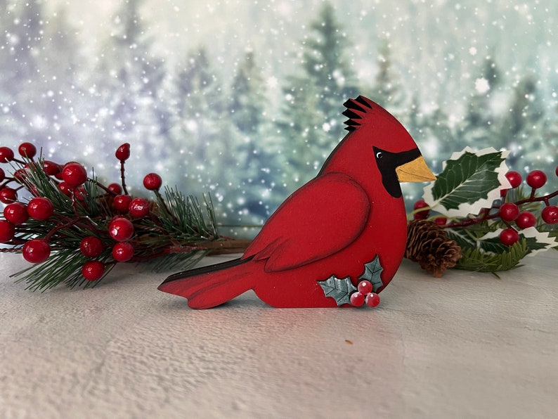 Pode incluir: Uma figura de p&aacute;ssaro cardeal vermelho com um olho e bico pretos, pousada em uma superf&iacute;cie nevada com um ramo de azevinho e bagas vermelhas.