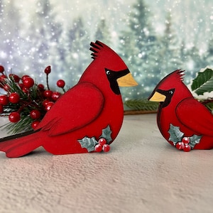 Pode incluir: Dois ornamentos de p&aacute;ssaros cardeais de madeira vermelha com azevinho e bagas. Os cardeais est&atilde;o em p&eacute; sobre uma superf&iacute;cie branca.