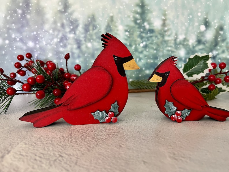 Pode incluir: Dois ornamentos de p&aacute;ssaros cardeais de madeira vermelha com azevinho e bagas. Os cardeais est&atilde;o em p&eacute; sobre uma superf&iacute;cie branca.