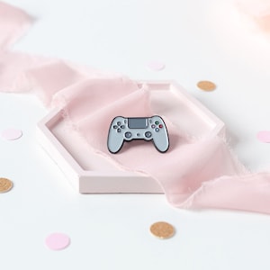 May include: A silver-coloured video game controller pin on a pink fabric background. The pin is a replica of a popular gaming console controller.