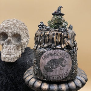 May include: A green frog wearing a witch's hat sits atop a small, round, black and gold container. The container has a label that reads "Crow's Feet Apothecary Organic Wart Enhancer".