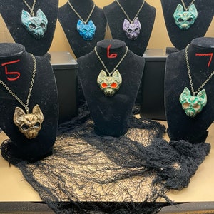 May include: Seven necklaces with different colored bat skull pendants. The pendants are made of ceramic and have intricate details. The necklaces are displayed on black velvet stands.