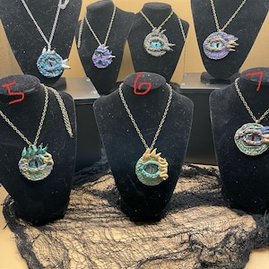 Può includere: Sette collane con ciondoli a forma di occhio di drago. I ciondoli sono in metallo e hanno un design testurizzato e squamoso. Gli occhi sono blu e verdi con accenti dorati. Le collane sono esposte su supporti in velluto nero.