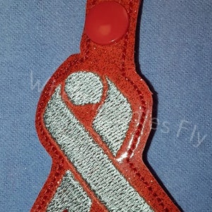 May include: Red glitter keychain with a silver ribbon and a red button. The ribbon is in the shape of an awareness ribbon.