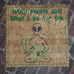 Pode incluir: Um descanso de copo de cortiça com costuras verdes ao redor das bordas. O descanso de copo apresenta um alienígena bordado segurando um objeto verde. O texto "When people ask what I do for fun" é bordado em verde no descanso de copo.