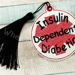 Puede incluir: Círculo de tela blanca con un símbolo médico rojo y el texto "Insulin Dependent Diabetic" en letras negras. Un borla negra está unida al círculo.