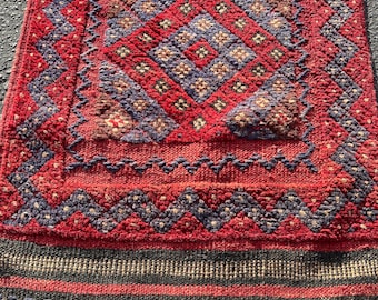 Puede incluir: Una alfombra roja, azul y negra con un diseño geométrico. La alfombra tiene un borde de flecos.