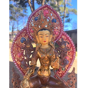 May include: A detailed statue of a seated deity with a golden complexion, adorned with a jeweled crown and necklace. The statue features intricate carvings and is set against a backdrop of a red and gold decorative element. The overall aesthetic is ornate and spiritual.
