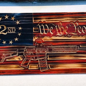 以下が含まれることがあります： 焦げ付いた木製のアメリカ国旗。旗には「We the People」と「2nd」の文字が刻まれている。赤と白のストライプの上にライフルのシルエットが重ねられている。