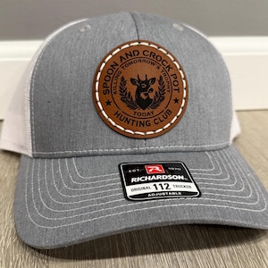 Pode incluir: Um boné de caminhoneiro cinza e branco com um remendo de couro marrom que diz "Spoon and Crock Pot Hunting Club" com uma silhueta de veado e o texto "Killing Tomorrow's Trophy Today". O boné é ajustável e tem um logotipo Richardson.