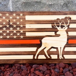 May include: Wooden American flag with a red stripe and a deer silhouette. The flag is made of stained wood with a natural wood finish.