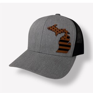 Peut inclure: Un chapeau de camionneur gris et noir avec un patch en cuir brun en forme de l'état du Michigan. Le patch présente un motif de drapeau américain noir et blanc.