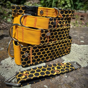 May include: A collection of dog collars and a matching lead. The collars display a black honeycomb pattern with yellow details and bee illustrations. Each collar has a metal D-ring and a black plastic buckle. The lead has a similar design.